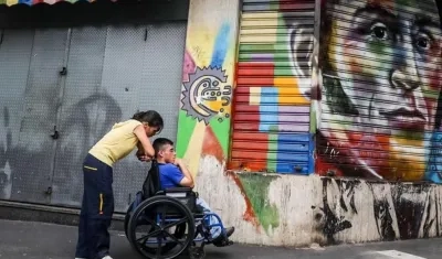 Una mujer empuja a un hombre en silla de ruedas frente a un graffiti del héroe nacional Simón Bolívar.